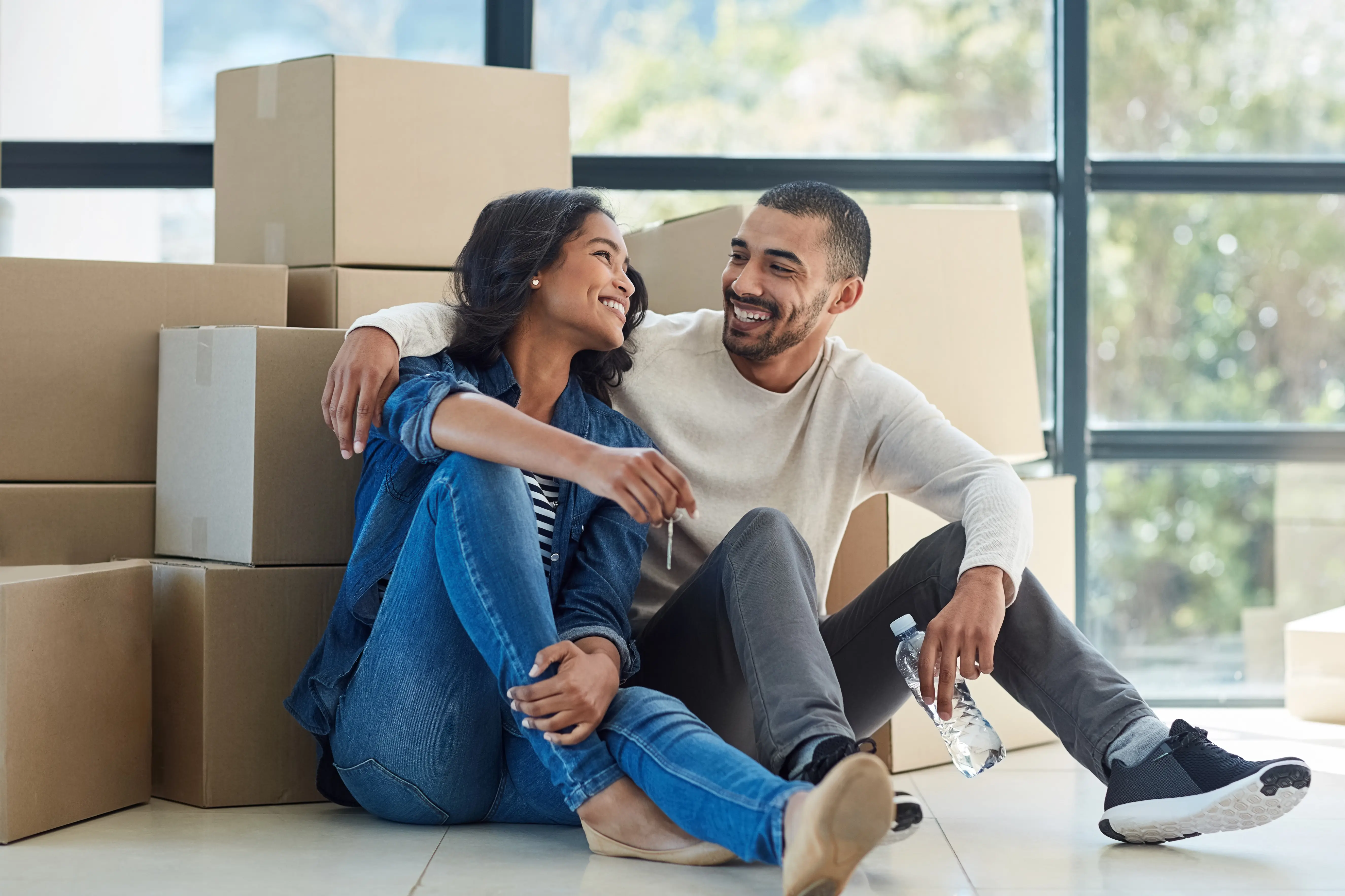 Happy couple sitting on the floor surrounded by moving boxes, smiling at each other